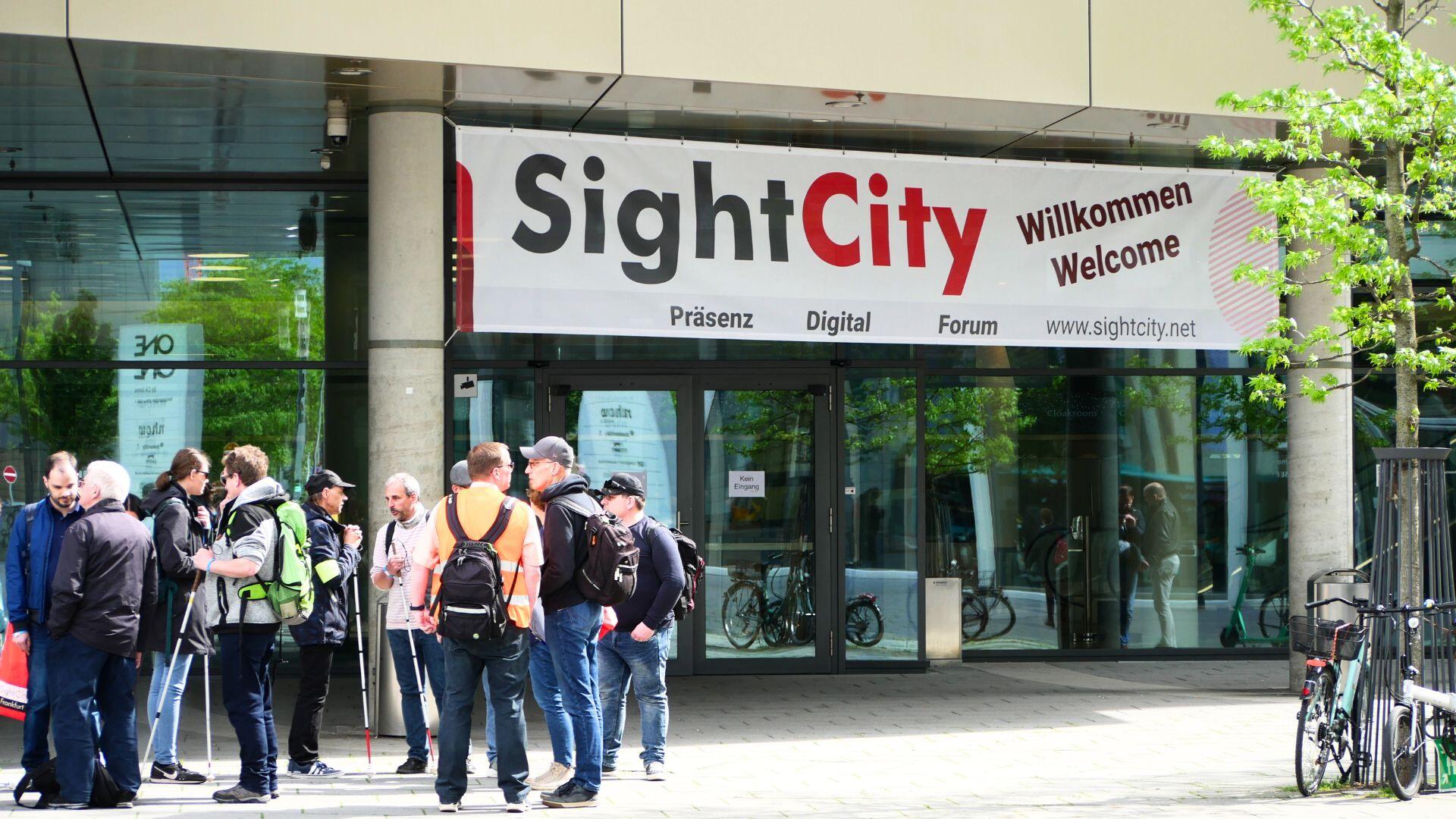 links steht eine Gruppe von Menschen, tw. mit Blindenstock vor dem Eingang zum Kap Europa. Über dem Eingang hängt ein Banner mit dem SightCity Logo und den Worten "Willkommen, Welcome"