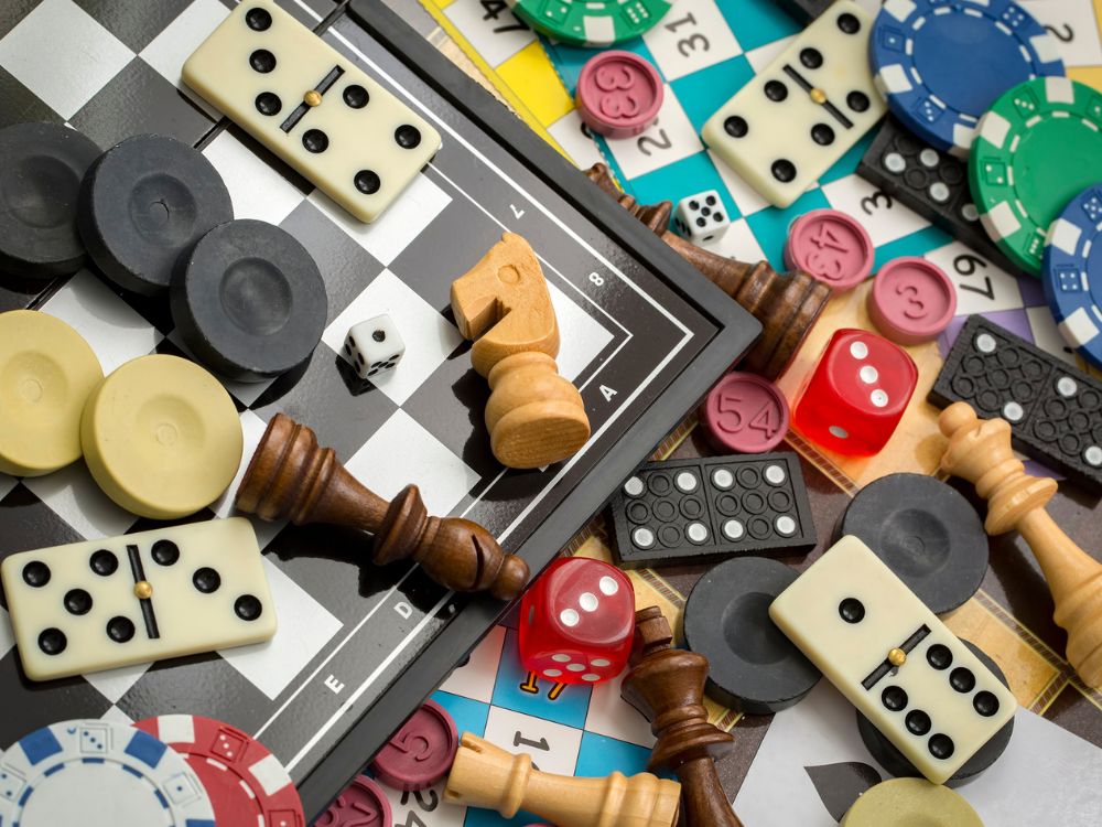 Eine bunte Sammlung verschiedener Spielmaterialien auf einem Tisch. Zu sehen sind Dominosteine, Schachfiguren, Würfel in verschiedenen Farben (rot, weiß), Spielchips in blau und grün, nummerierte Spielmarken und ein Schachbrett. Die Spielelemente überlappen sich und zeigen die Vielfalt analoger Gesellschaftsspiele.