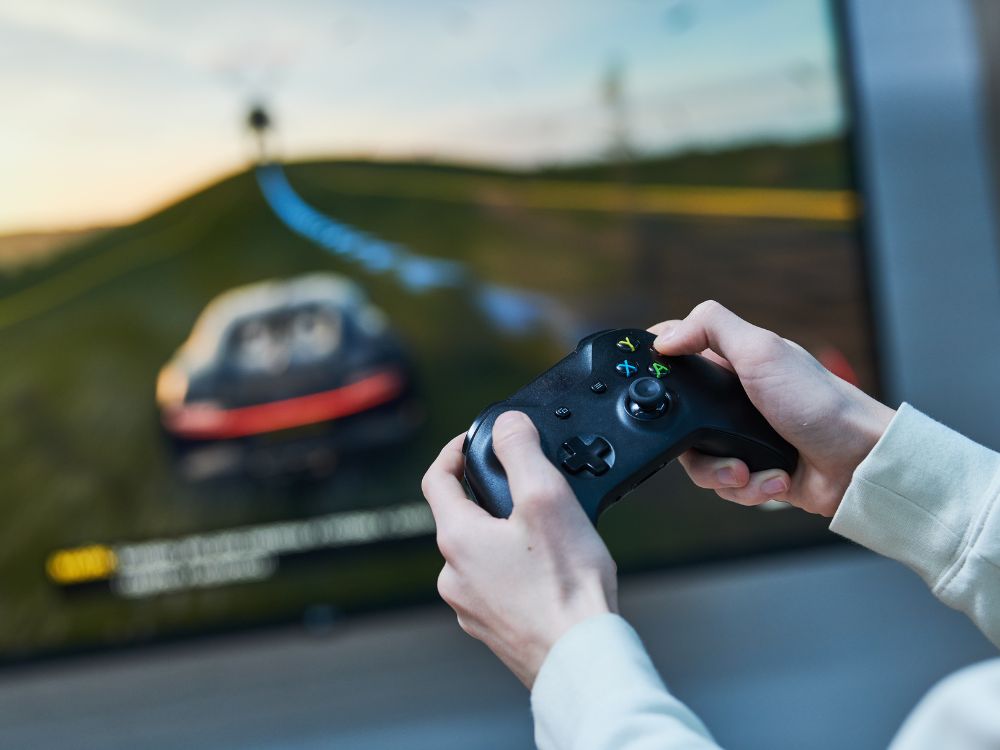 Nahaufnahme einer Person mit einem schwarzen Xbox-Controller in den Händen. Die Person spielt ein Rennspiel, das auf einem Bildschirm zu sehen ist. Auf dem Bildschirm ist ein verschwommenes Bild eines Rennwagens auf einer Strecke erkennbar. Die Hände tragen einen weißen Pullover und halten den Controller in Spielposition.
