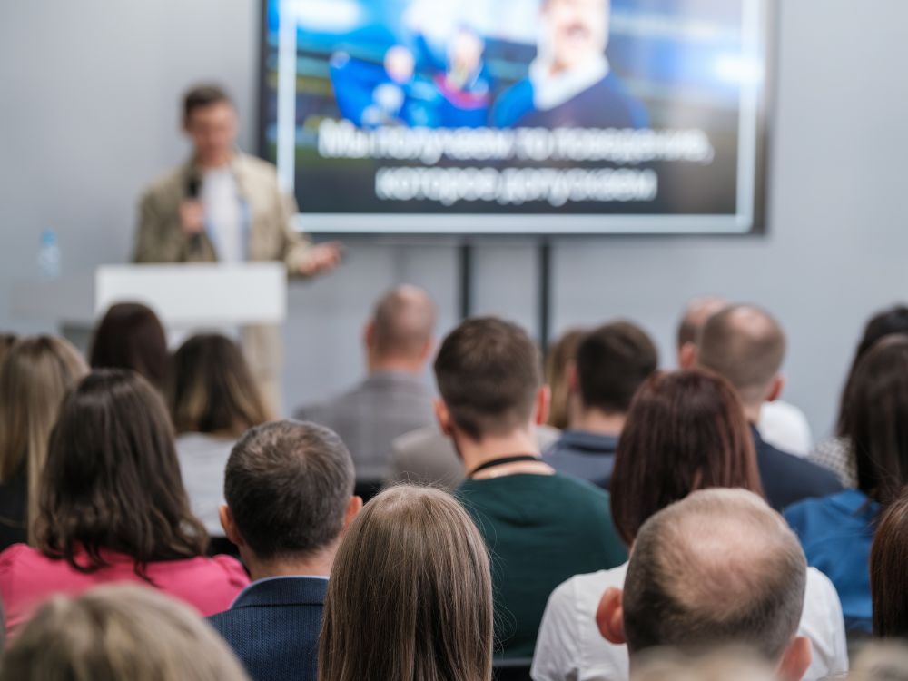 Ein Vortragender steht an einem weißen Rednerpult und präsentiert vor einem Publikum. Im Hintergrund ist ein großer Bildschirm mit einer Präsentation zu sehen. Das Publikum sitzt mit dem Rücken zur Kamera und besteht aus zahlreichen Zuhörern unterschiedlichen Alters. Die Szene zeigt eine Informationsveranstaltung oder einen Workshop zum Thema Gaming.